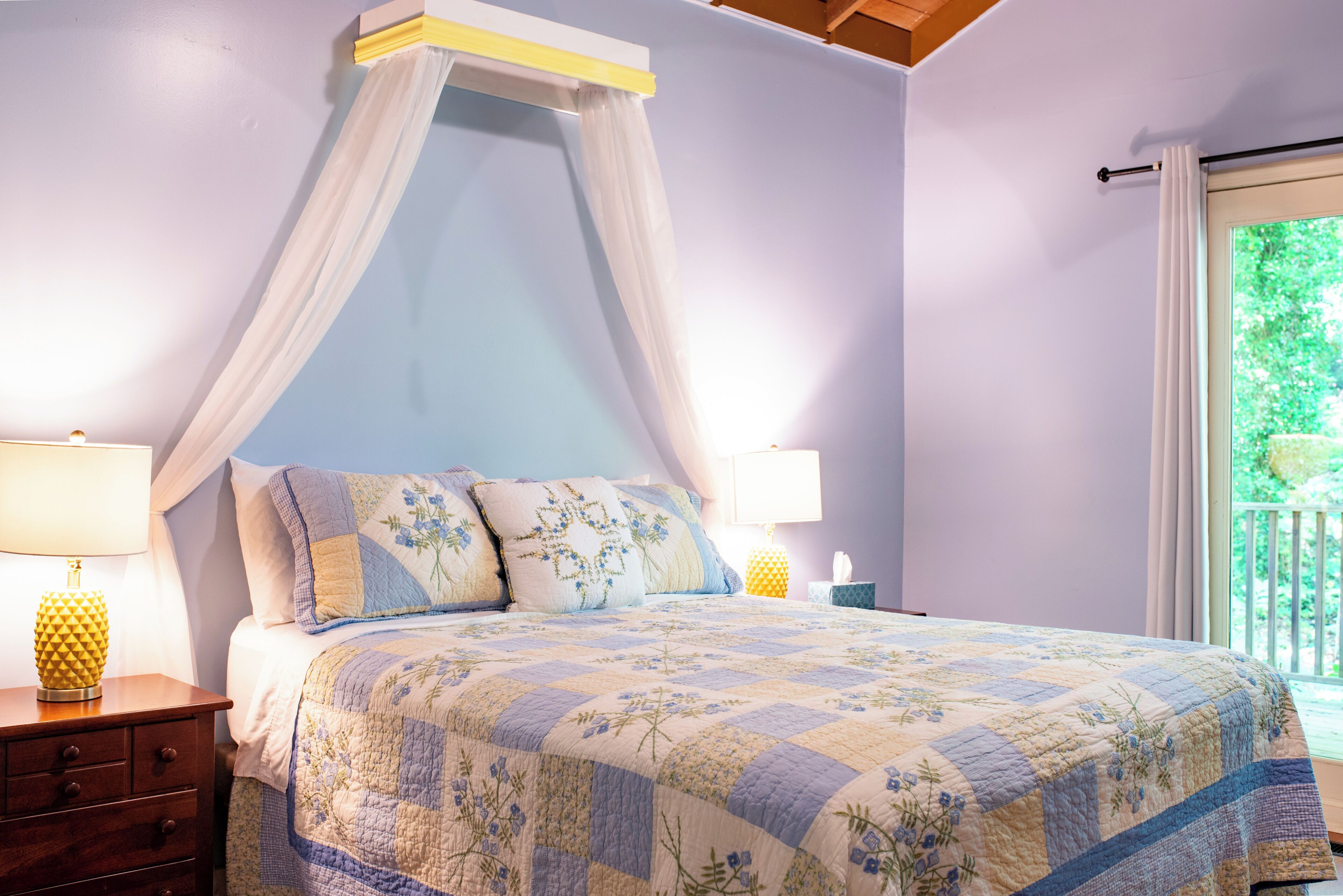 A bedroom with a canopy bed, bedside lamps, a nightstand, and a window with a view of greenery.