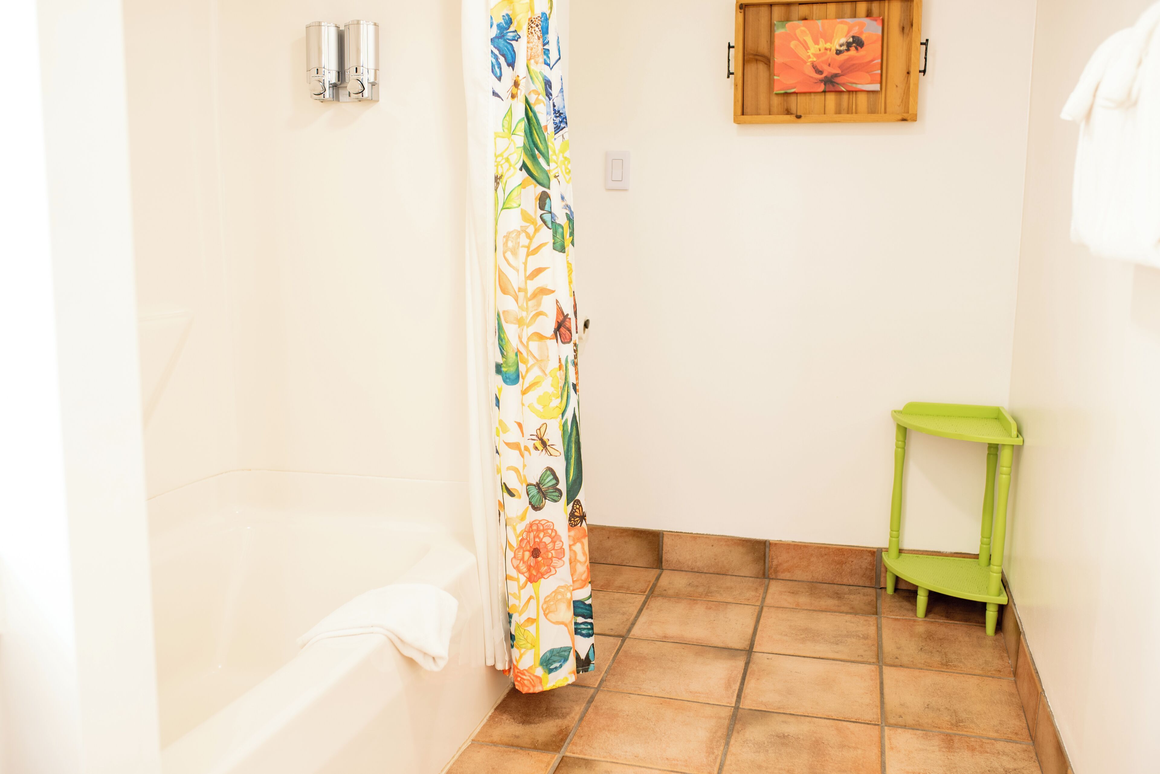 A bathroom with a bathtub, a shower curtain with a floral pattern, a green shelf, and a framed picture on the wall.