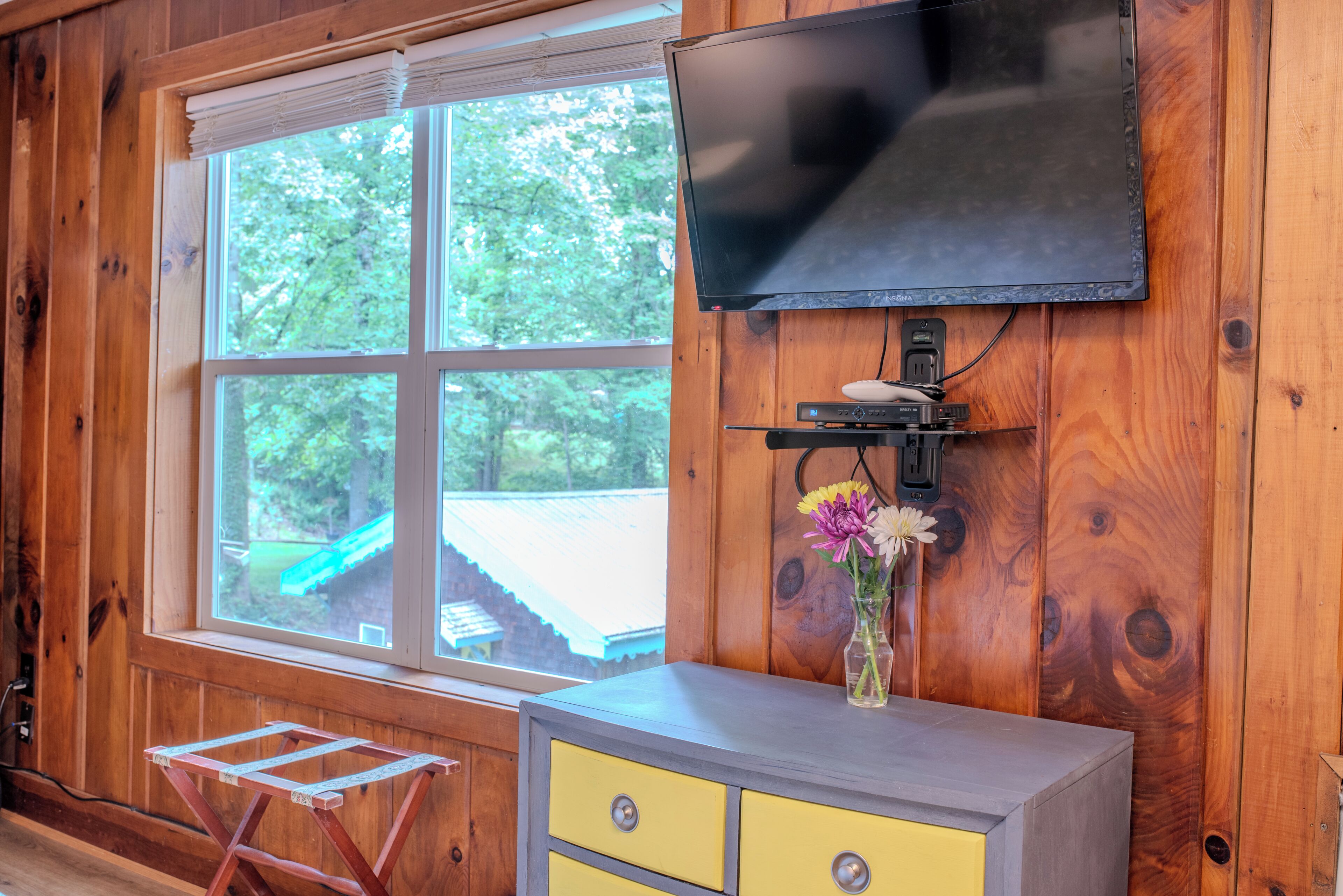 Ein Zimmer mit Holzwänden, an der Wand ist ein Fernseher angebracht, ein kleiner Tisch mit einem Blumenstrauß und ein Fenster mit Blick auf Grünfläche.