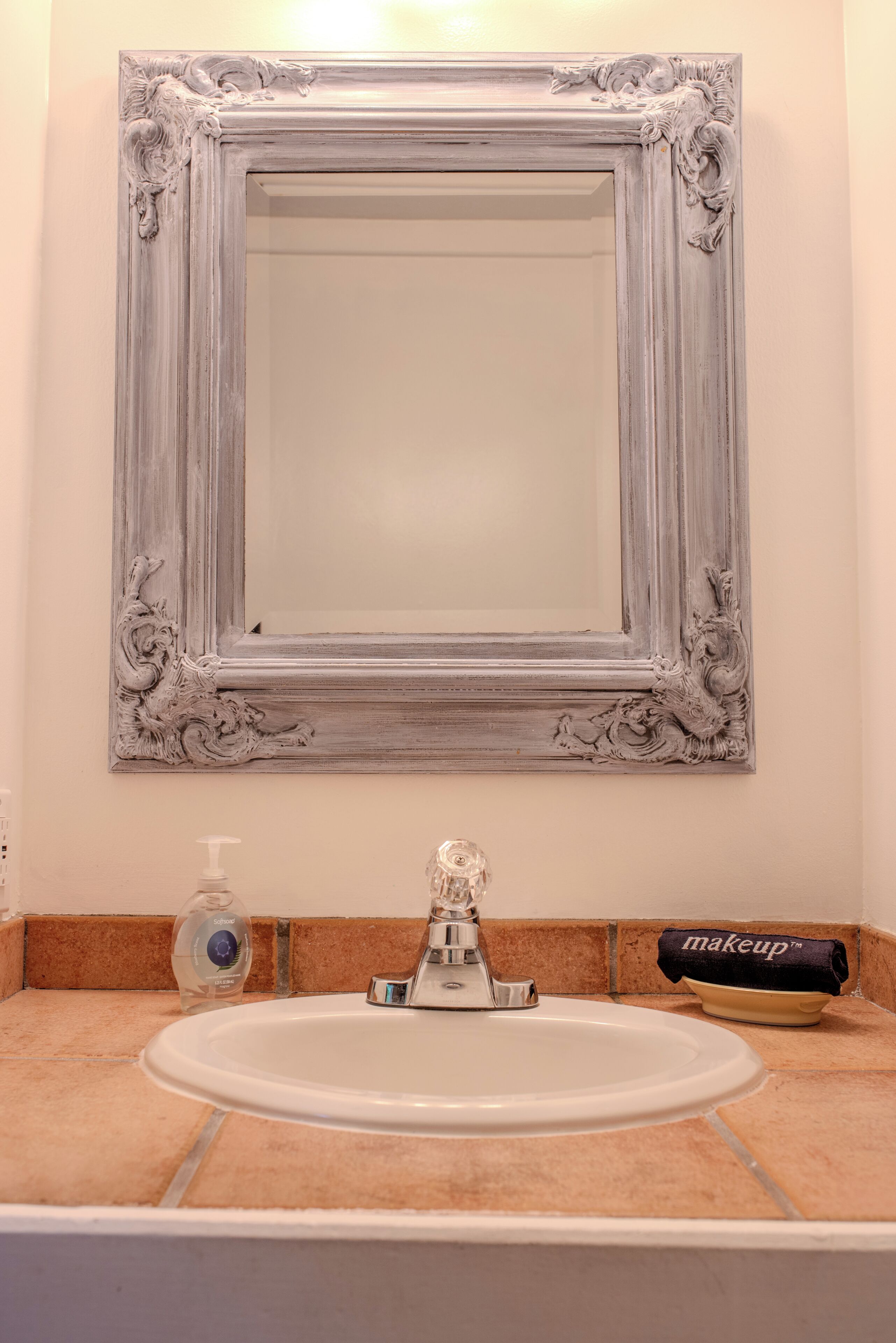 Une salle de bain avec un lavabo blanc, un miroir doté d’un cadre orné et une serviette pour se maquiller posée sur le comptoir.