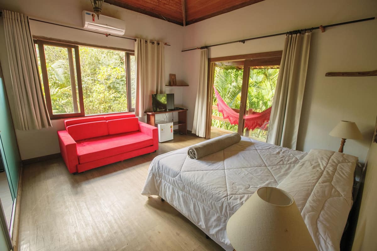 A bedroom with a bed, a red sofa, a TV, and a window with a view of greenery.