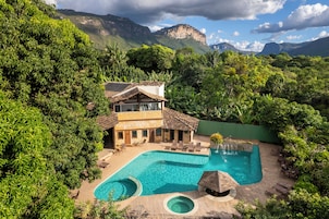 Un edificio de dos pisos con piscina rodeada de exuberante vegetación y montañas al fondo.