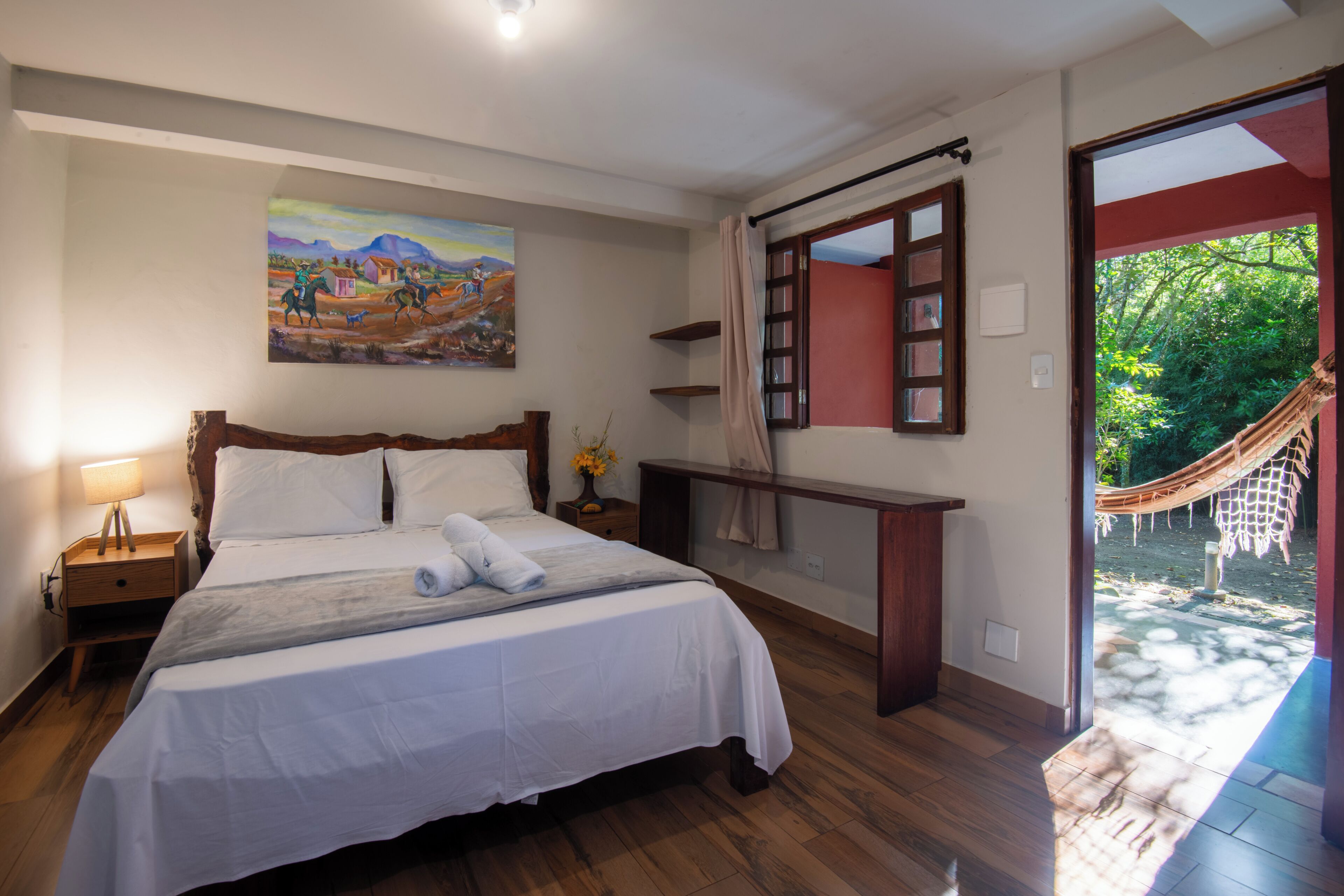 A bedroom with a bed, a nightstand, a painting, a window with a view of greenery, and a hammock outside.