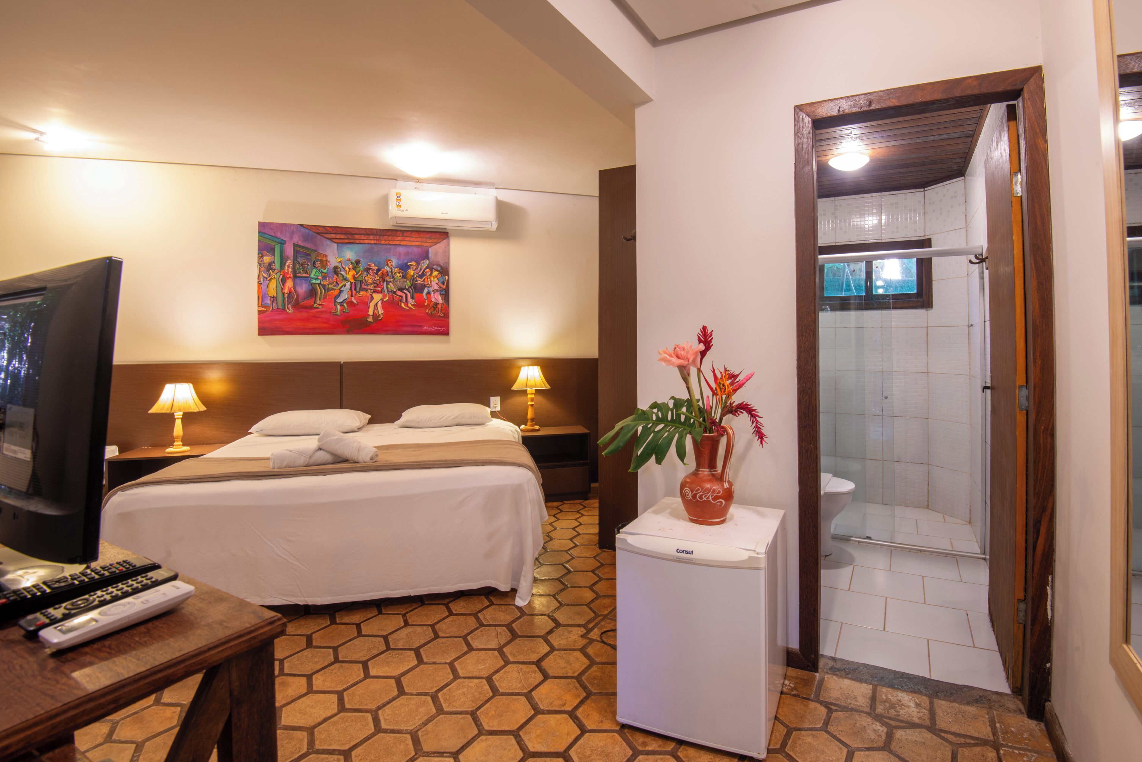 Une chambre d’hôtel comprenant un lit, des lampes de chevet, un téléviseur, un tableau au mur, un vase avec des fleurs et un espace douche.