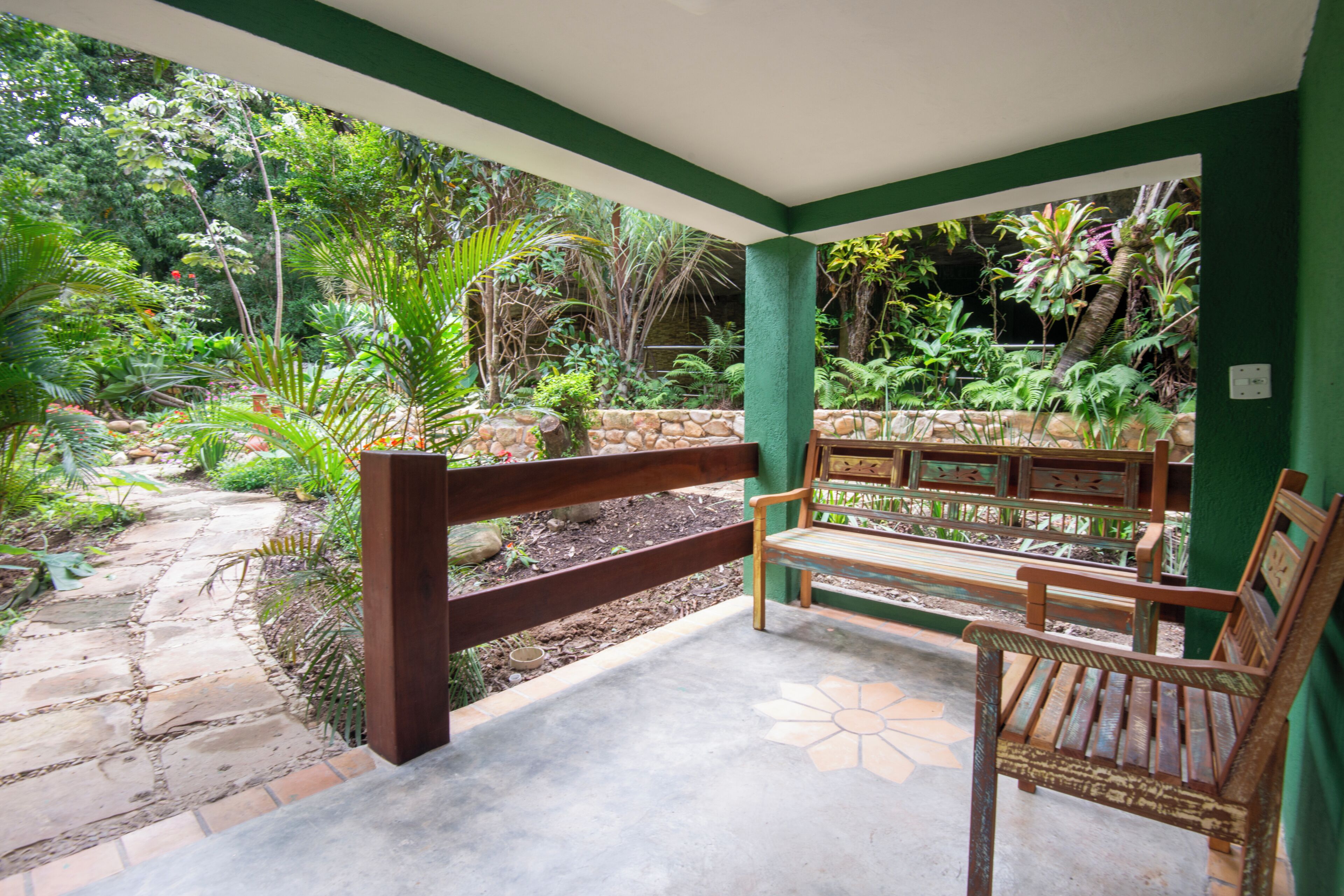 A covered outdoor seating area with benches, surrounded by lush greenery and a stone pathway.