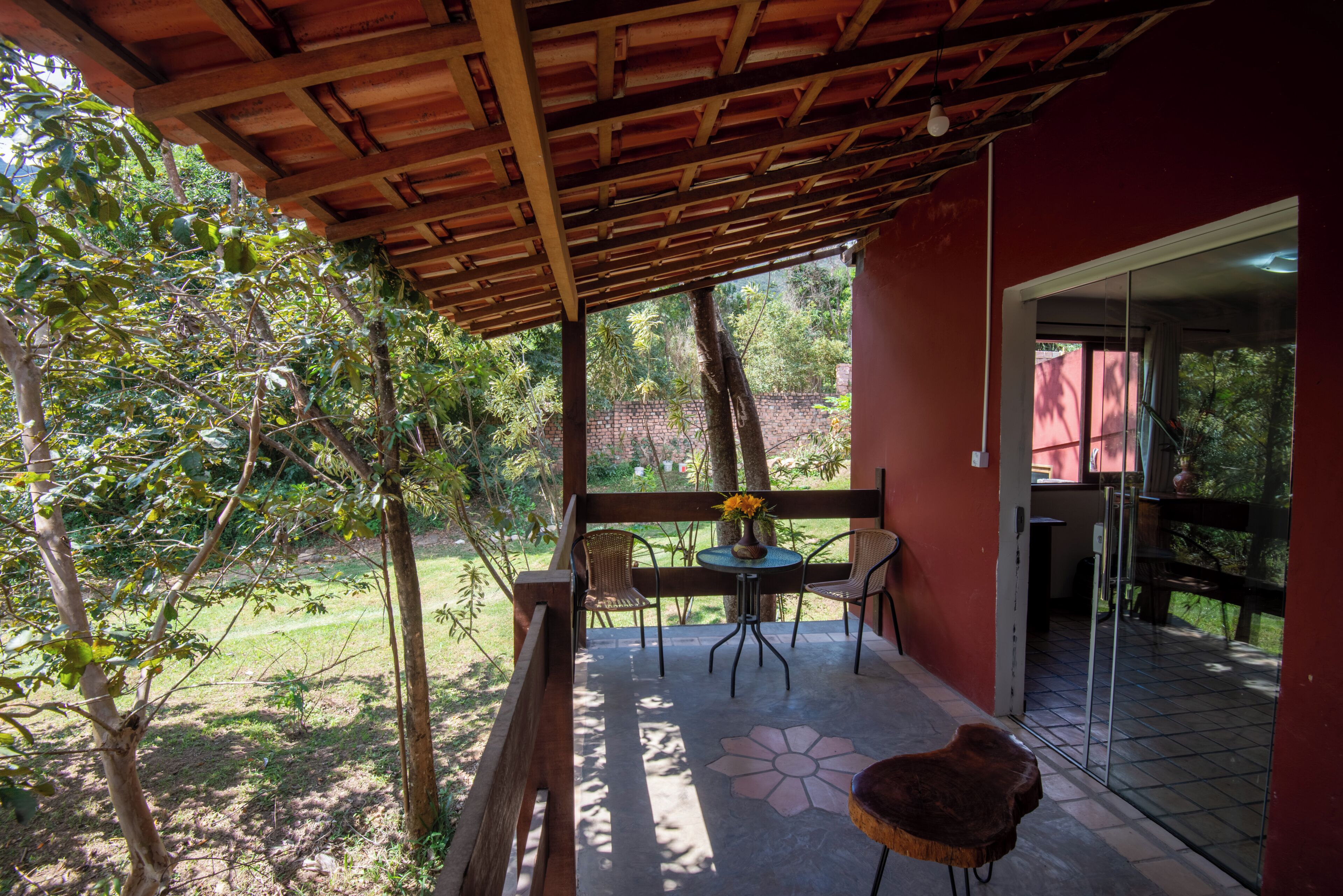 A covered patio with a table and chairs, a wooden bench, and a view of a garden and trees.