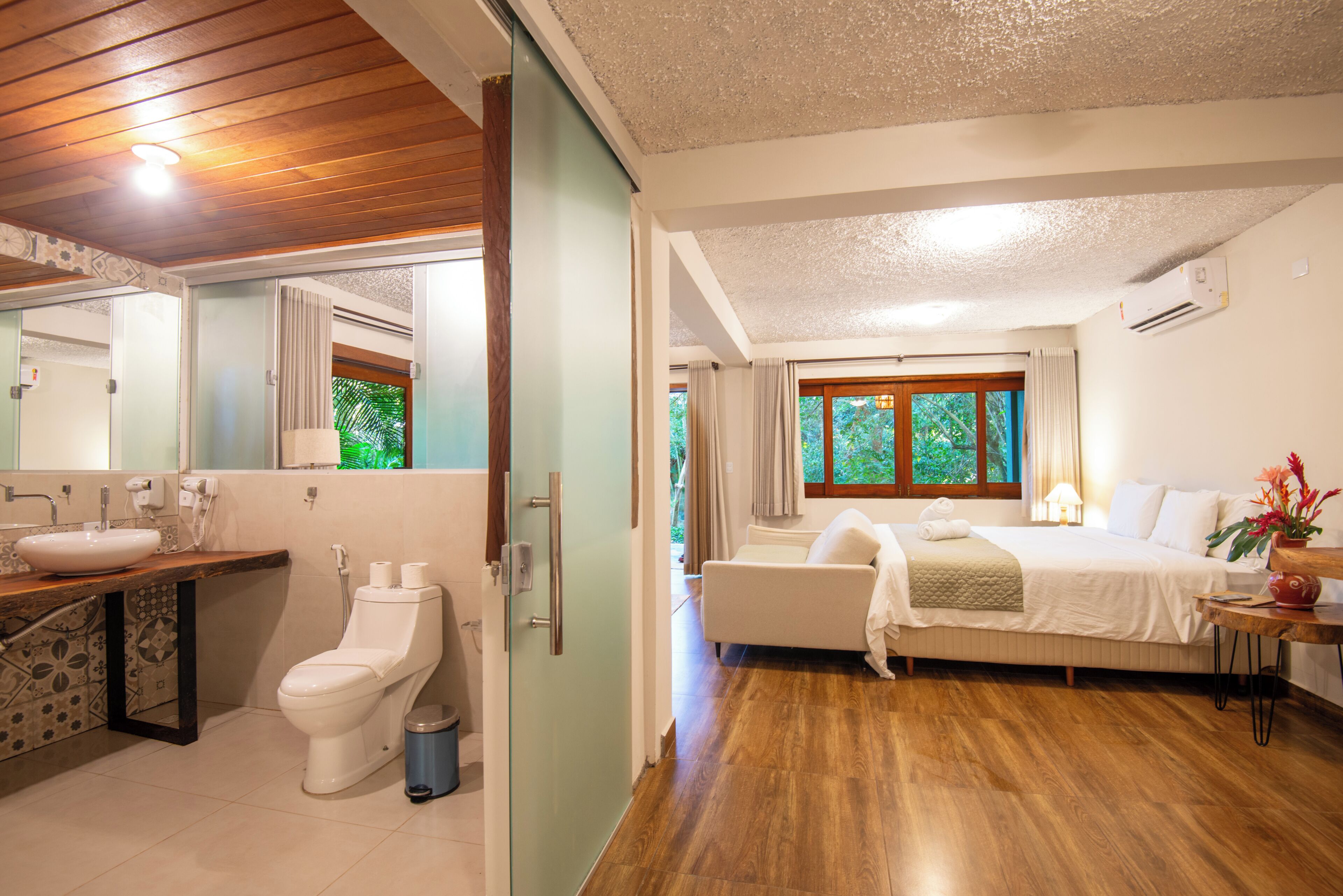 A hotel room with a bathroom, a bed, a bedside table, and a window with a view of trees.