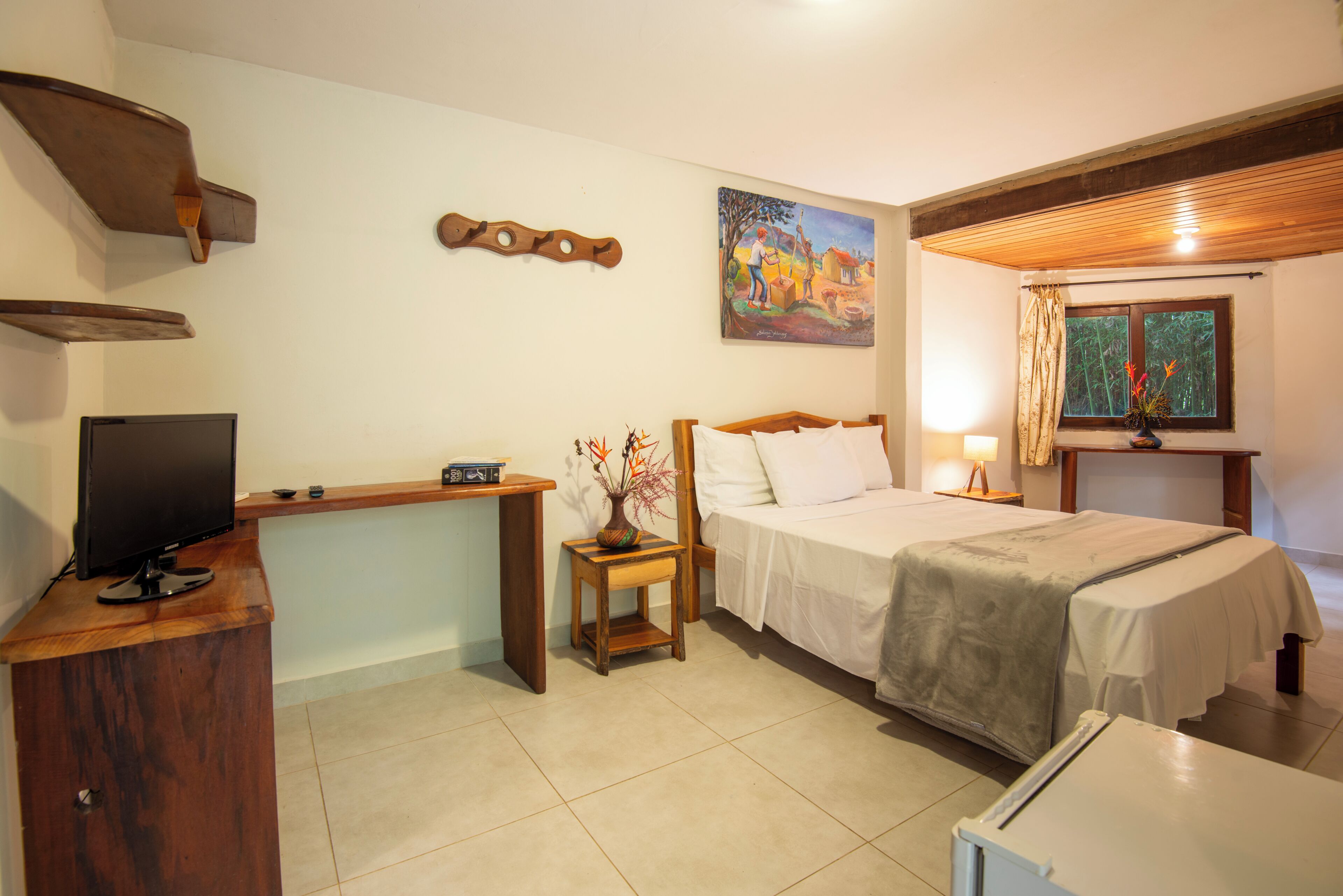 A hotel room with a bed, bedside table, desk with a television, a painting on the wall, and a window with curtains.