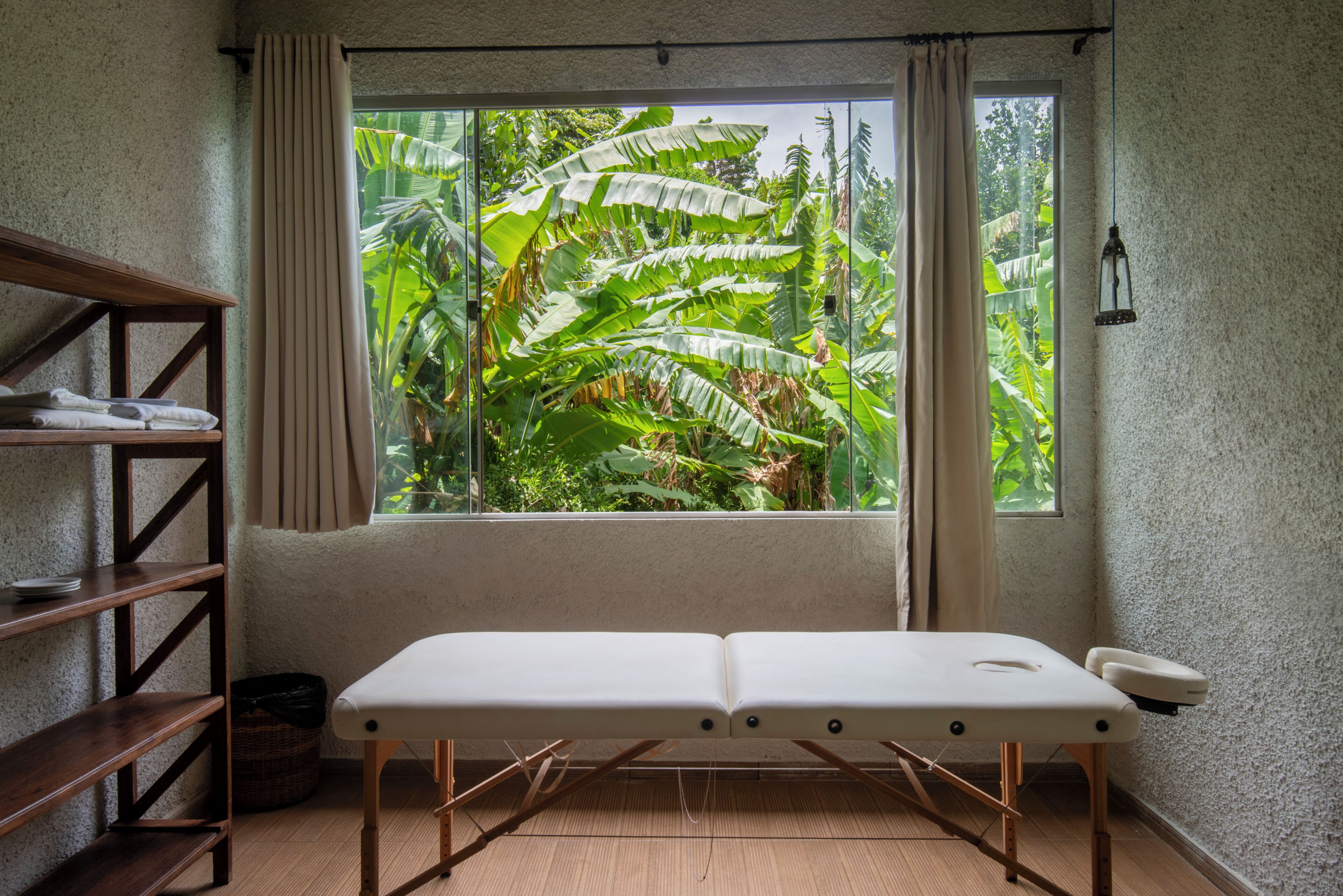 A room with a massage table, wooden shelves, a window with curtains, and a view of lush greenery outside.