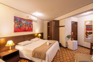 Quarto de hotel com cama, luminárias de cabeceira, um quadro, um espelho e uma pequena planta.