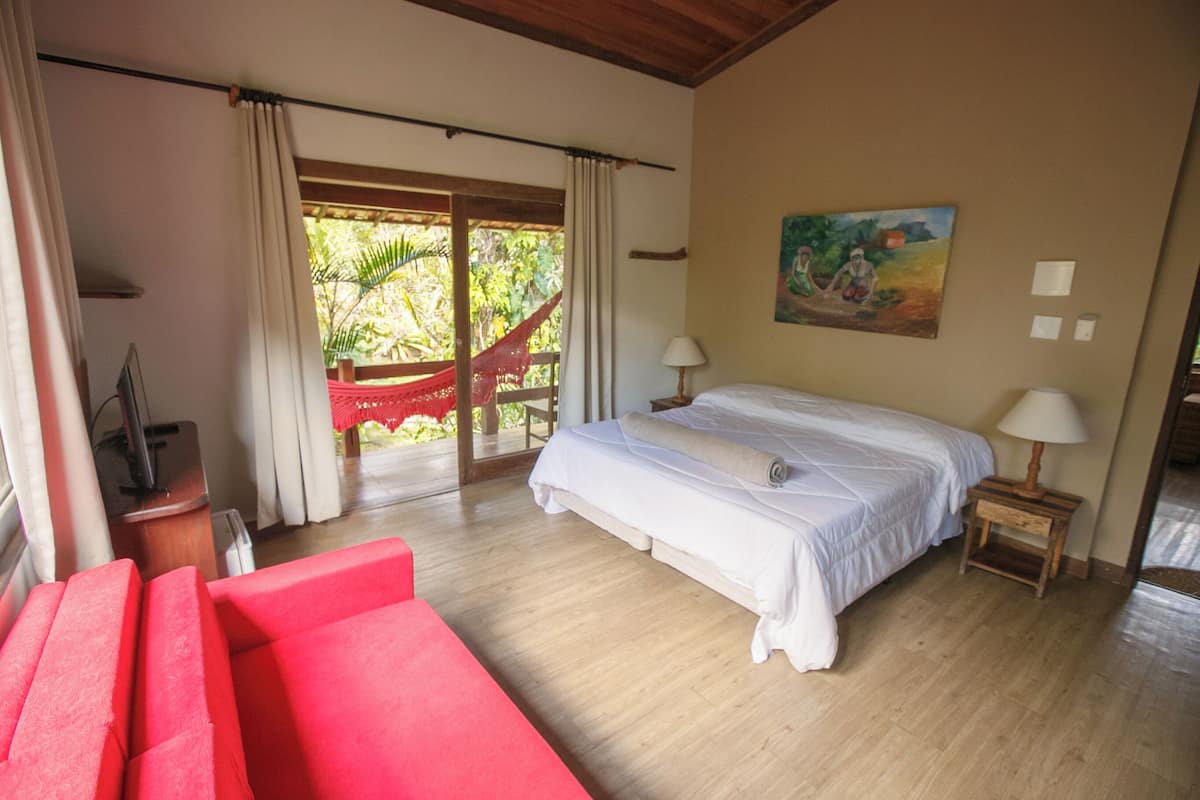 A bedroom with a large bed, two bedside tables, a red sofa, and a hammock outside the window.