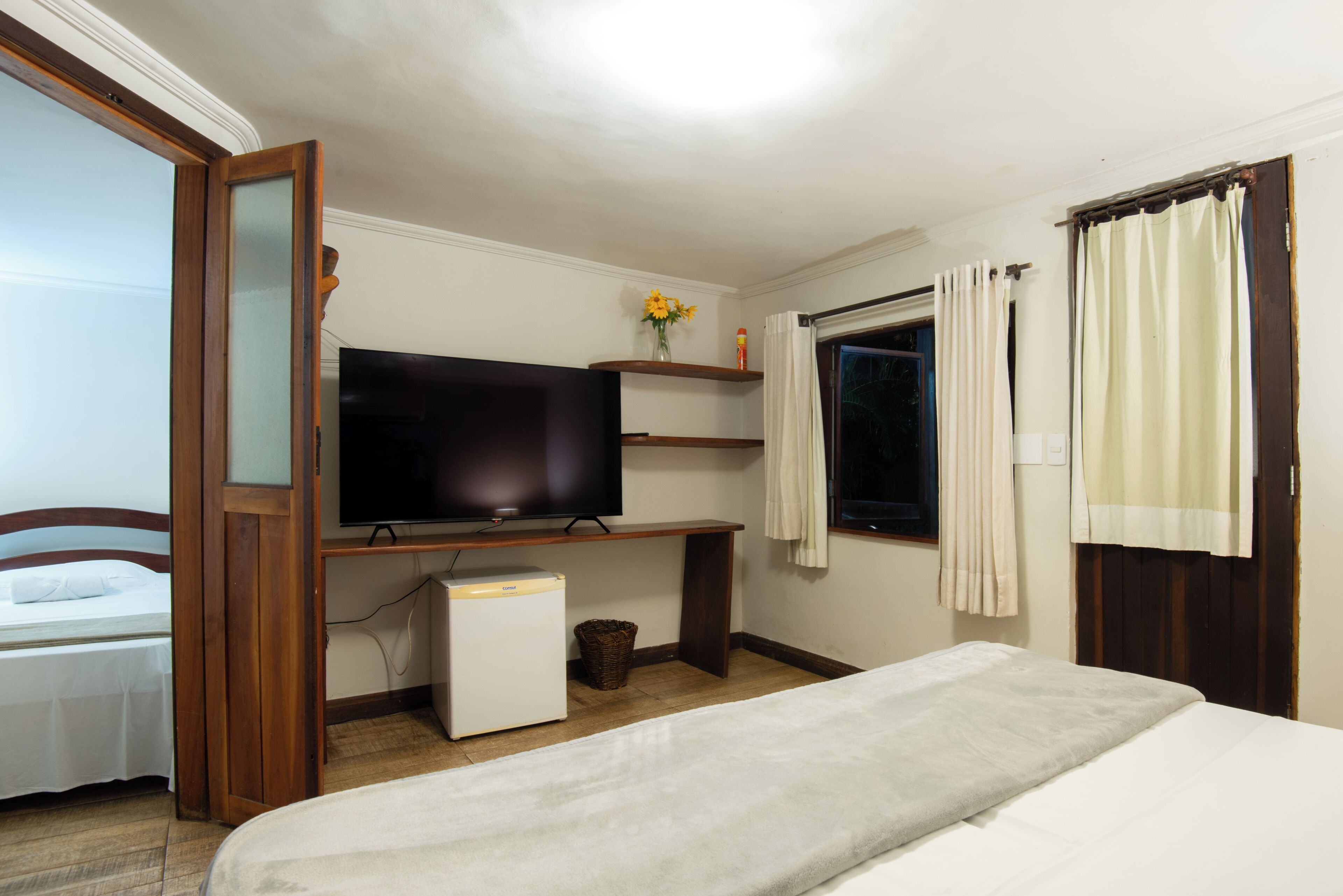A hotel room with a bed, a television, a small refrigerator, a window with curtains, and a wooden desk with a shelf above.