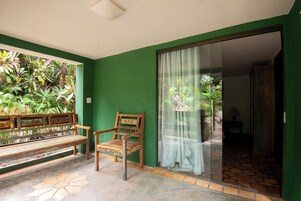 Una habitación con un banco y una silla, un ventanal corredero que da a un área exterior con plantas, y un piso de baldosas.