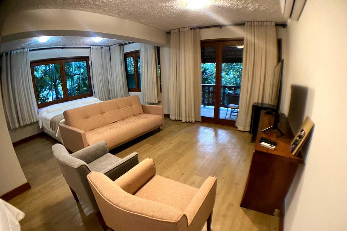A spacious living room with a sofa, armchair, and a TV on a wooden stand. Large windows with curtains offer a view of the outdoors.