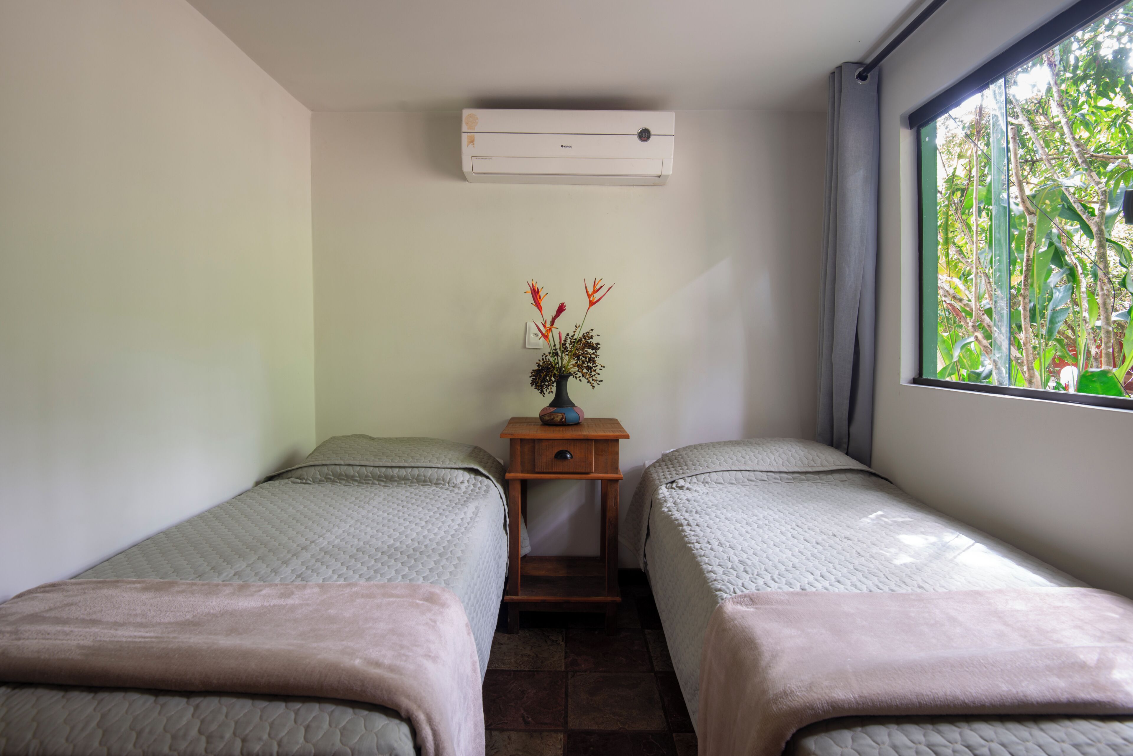 A room with two beds, a small wooden nightstand with a vase of flowers, and a window with a view of greenery.
