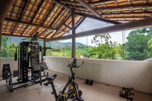 A gym with exercise equipment, a stationary bike, and a view of a mountainous landscape through large windows. - Pousada Lendas do Capão (Palmeiras)