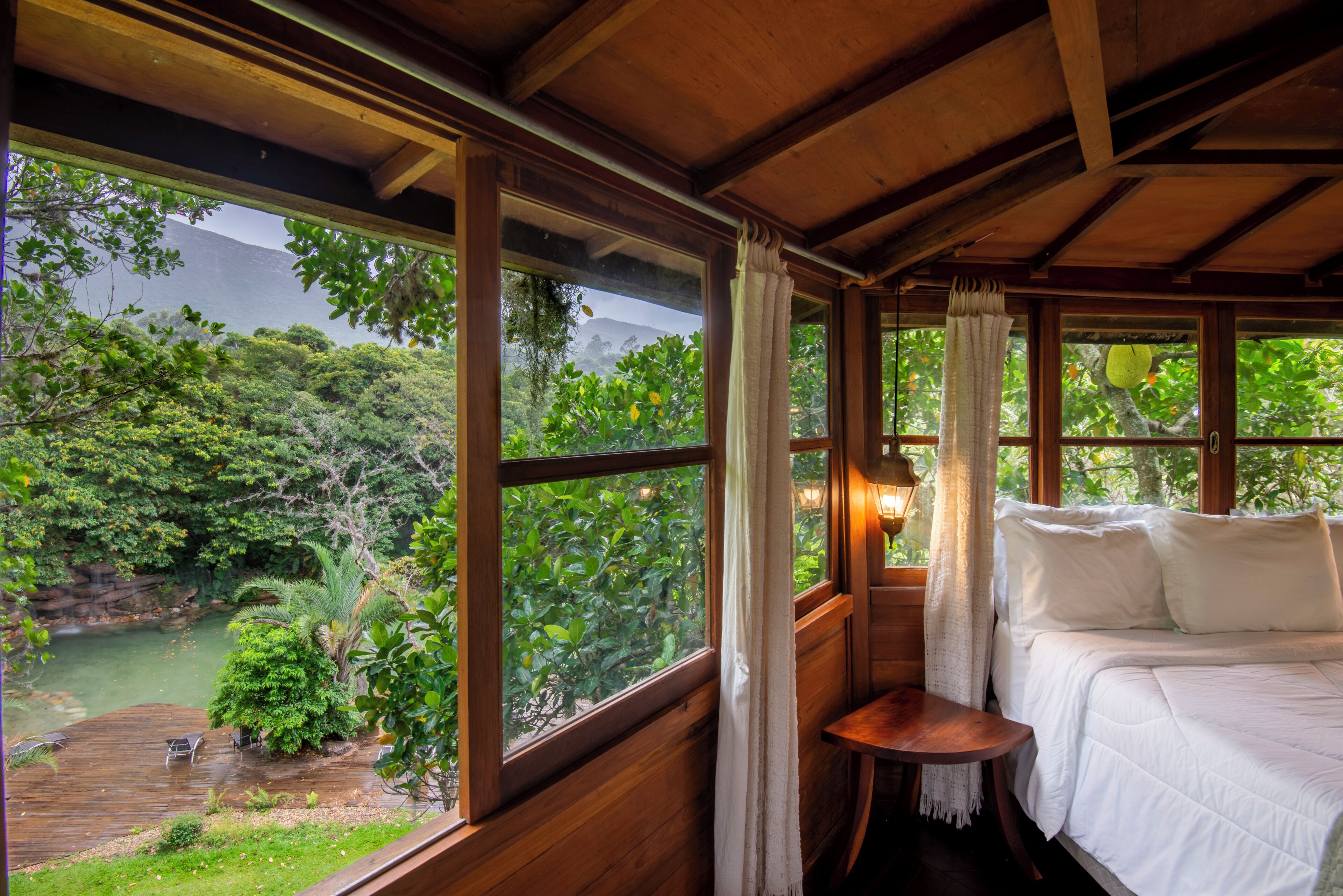 A wooden room with a bed, a small table, and large windows offering a view of a forest and a body of water.