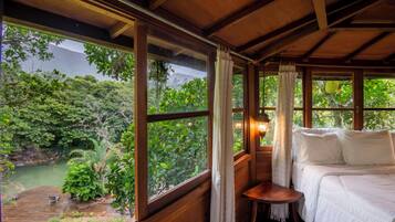 A wooden room with a bed, a small table, and large windows offering a view of a forest and a body of water.