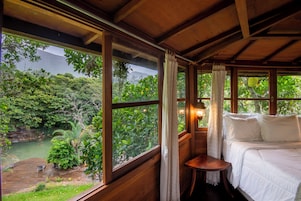 Quarto de madeira com cama, uma mesa pequena e grandes janelas que oferecem vista para uma floresta e um corpo d'água.