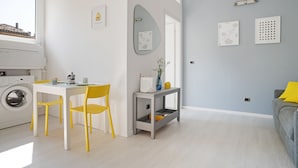 Una sala moderna con un sofá gris, un sillón amarillo, una mesita con un florero y una lavadora al fondo.