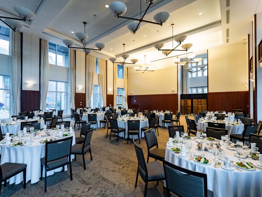 A large banquet hall with round tables set for dining, featuring chandeliers and large windows.