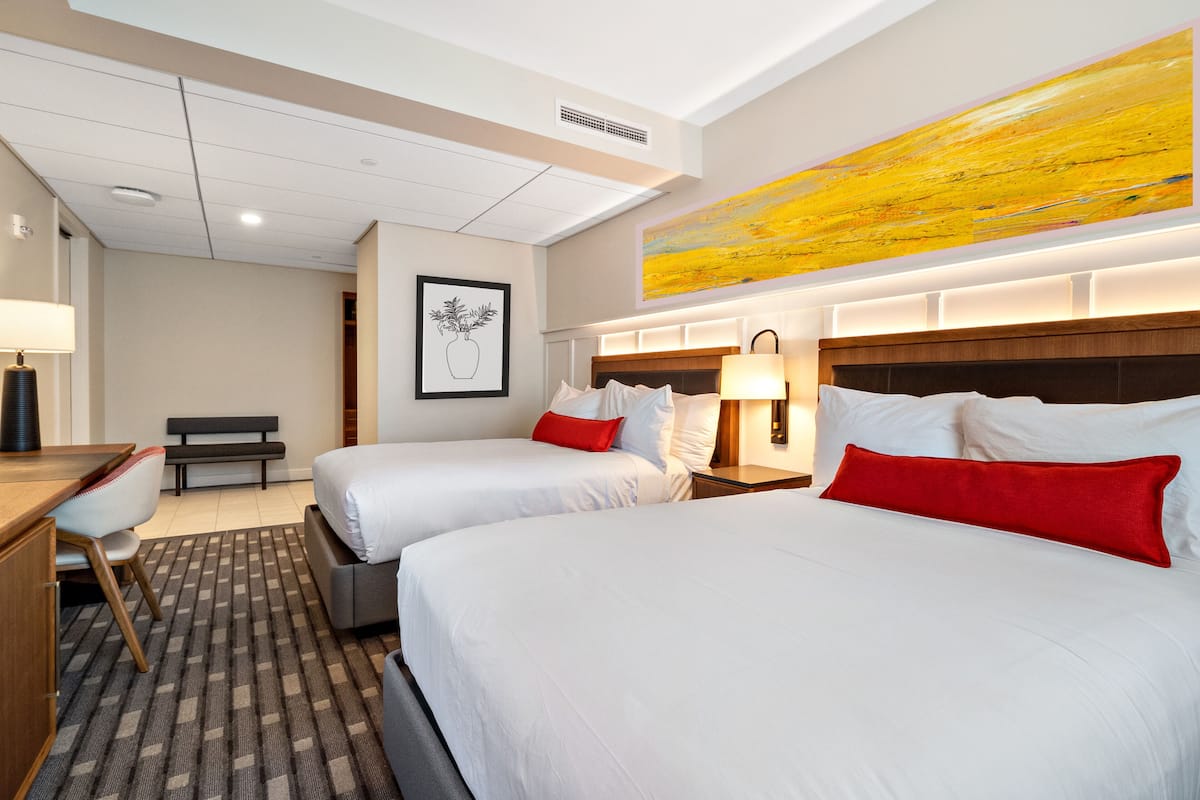 Une chambre d’hôtel avec deux lits, un bureau, une chaise, un banc et un grand tableau abstrait accroché au mur.