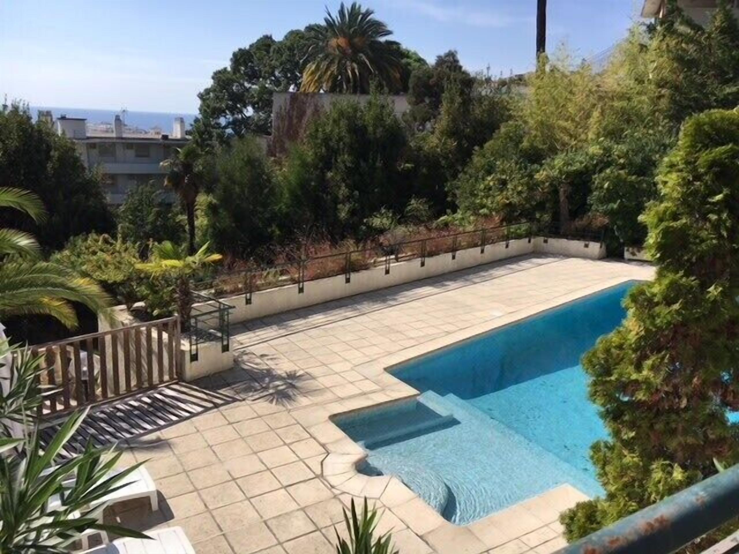 Une piscine rectangulaire avec une terrasse carrelée, entourée d’une végétation luxuriante et d’une rambarde en métal.