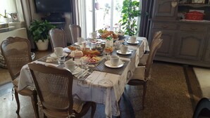 A dining table set with breakfast items, surrounded by wicker chairs, in a room with a television and plants. - Les iris Chambres D Hôtes (Niedermorschwihr)
