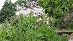 A garden with various plants and flowers, a white building with balconies, and a stone structure. - Les iris Chambres D Hôtes (Niedermorschwihr)