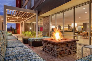 An outdoor seating area with a fire feature, cushioned seating, and a glass-enclosed dining area. - Home2 Suites by Hilton Glen Mills Chadds Ford, PA (Glen Mills)