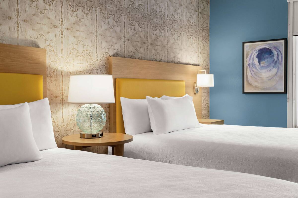 A hotel room with two beds, a round table with a lamp, and a wall with a patterned wallpaper and a painting.