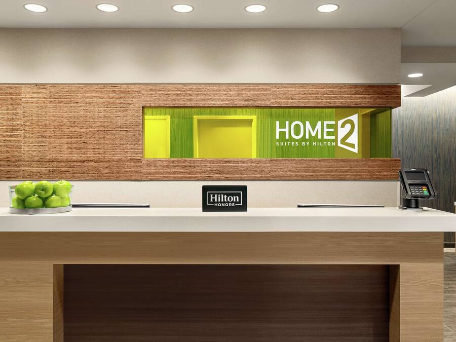 A hotel reception desk with a 'HOME2 Suites by Hilton' sign, a 'Hilton Honors' badge, and a digital payment terminal.