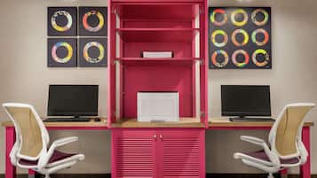 A workspace with two desks, each with a computer and a chair. There are two large wall-mounted displays and a pink cabinet with shelves.