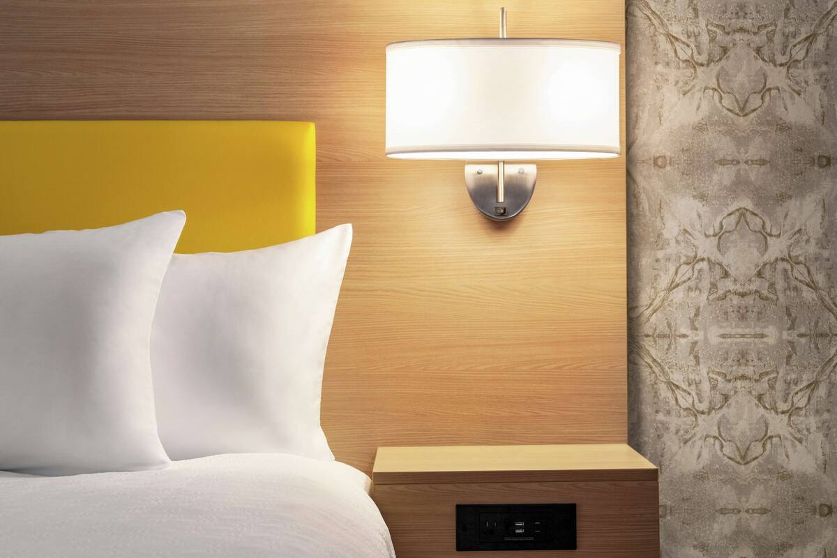 Una habitación de hotel con cabecera de madera, mesita de noche con lámpara y papel tapiz estampado.