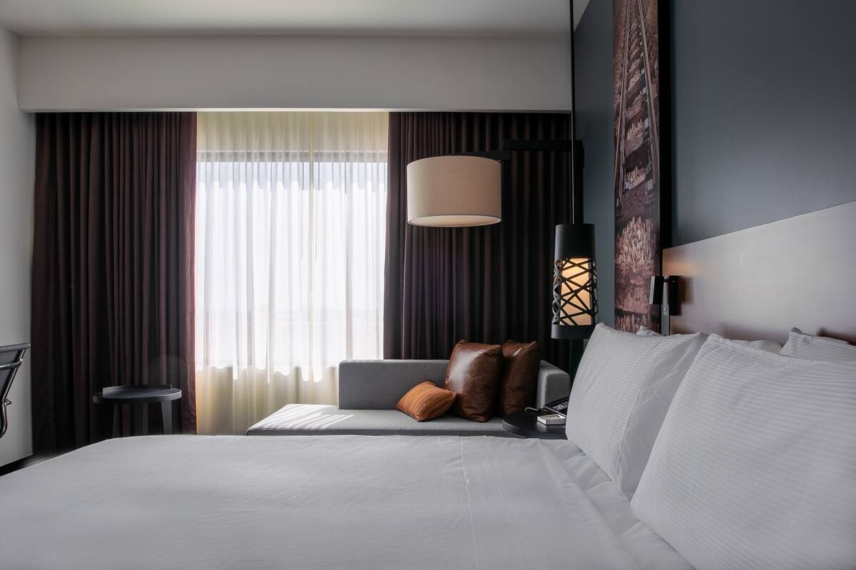 Quarto de hotel moderno com uma cama grande, uma mesa de cabeceira, um abajur e vista para o exterior pela janela.
