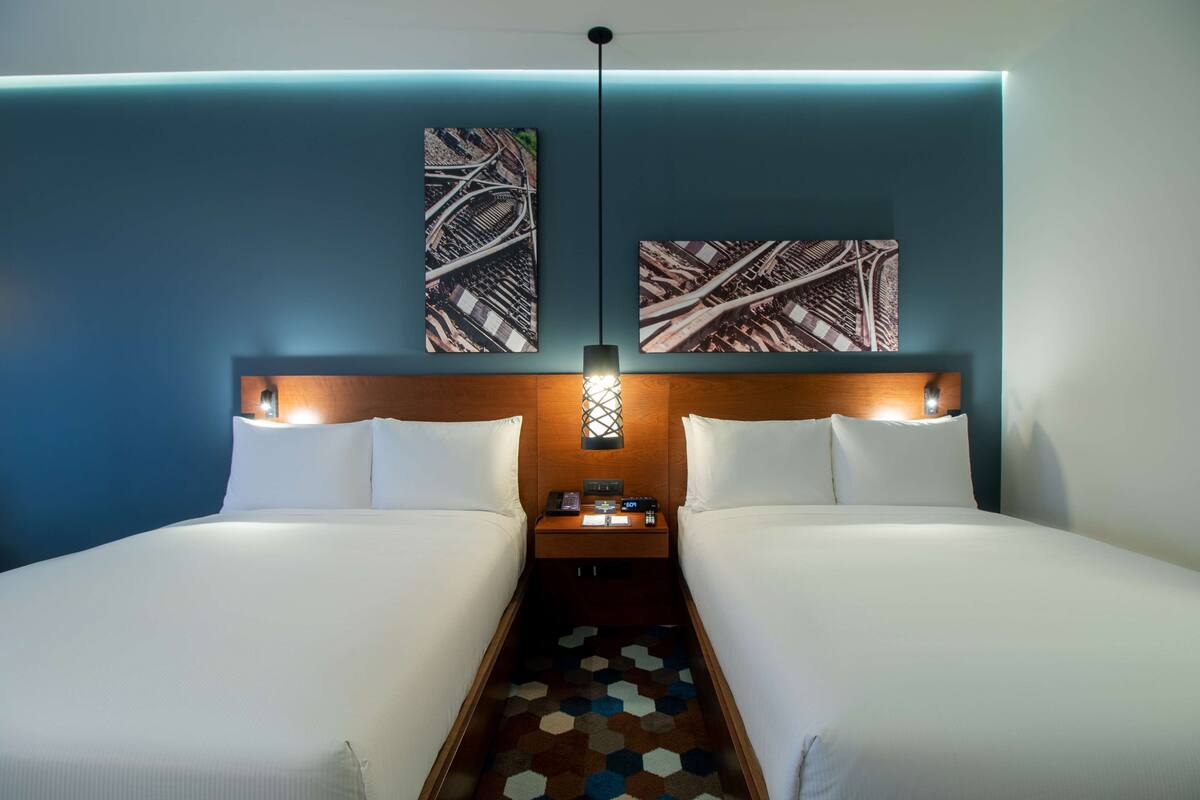 Quarto de hotel com duas camas, cabeceira de madeira, mesa de cabeceira com abajur e duas obras de arte penduradas na parede.