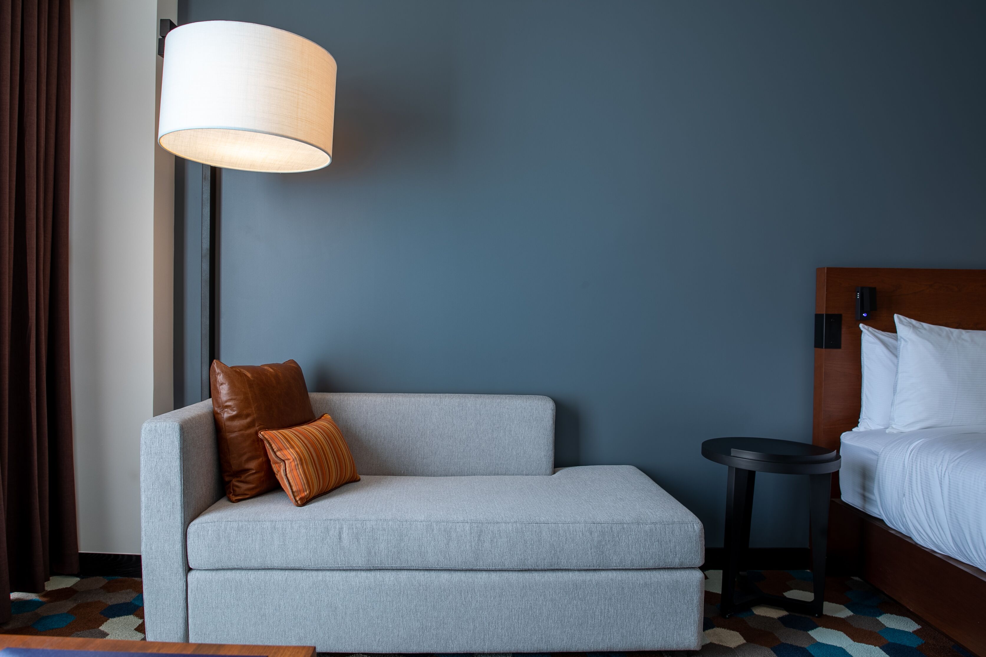 Habitación de hotel con un sofá gris, una cama con cabecera de madera, una mesita redonda pequeña y una lámpara de pie grande.