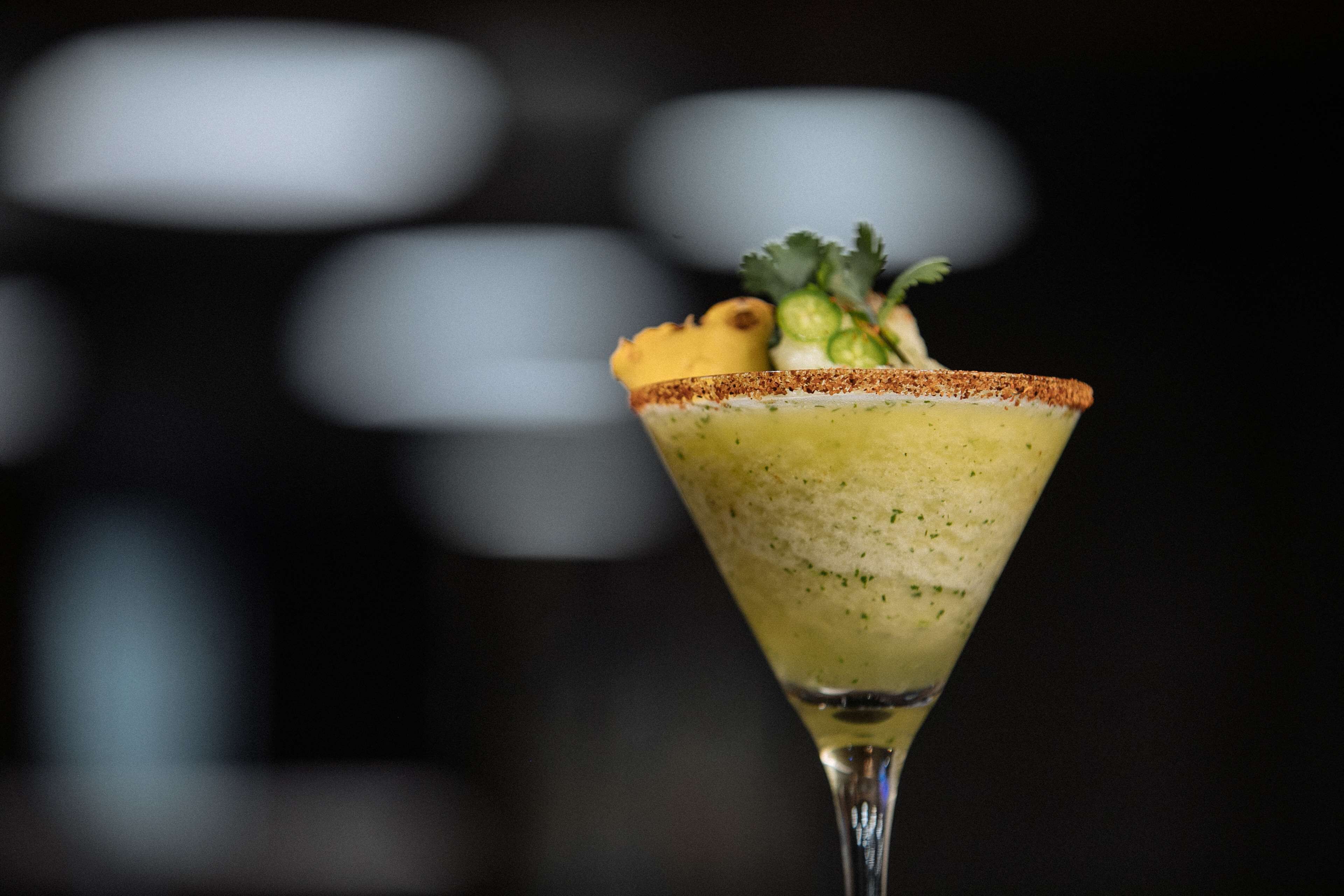 A cocktail in a martini glass with a salted rim, garnished with a slice of lime and a sprig of cilantro.