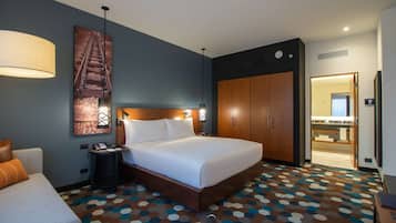 Habitación de hotel con una cama grande, un escritorio, una silla, una lámpara, un armario y un baño visible a través de una puerta abierta.