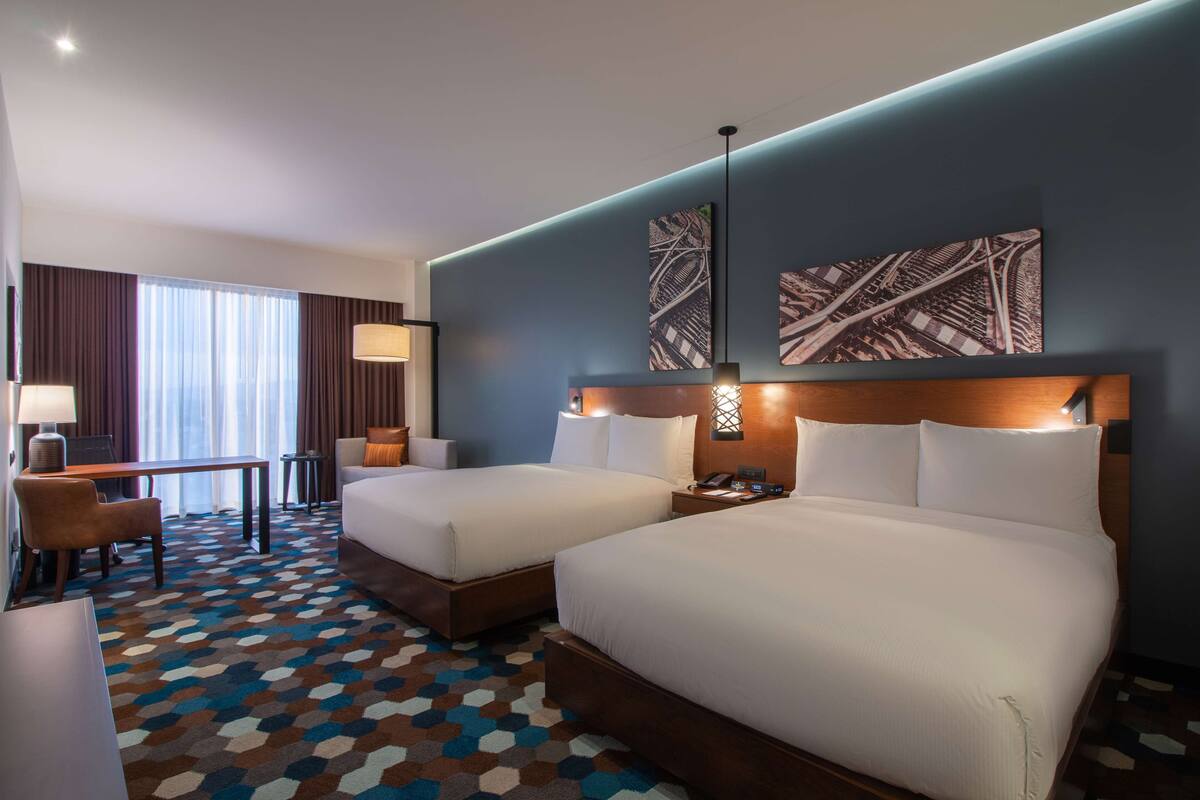 Quarto de hotel com duas camas, uma escrivaninha e uma cadeira. Há duas luminárias na parede e uma janela grande com cortinas.