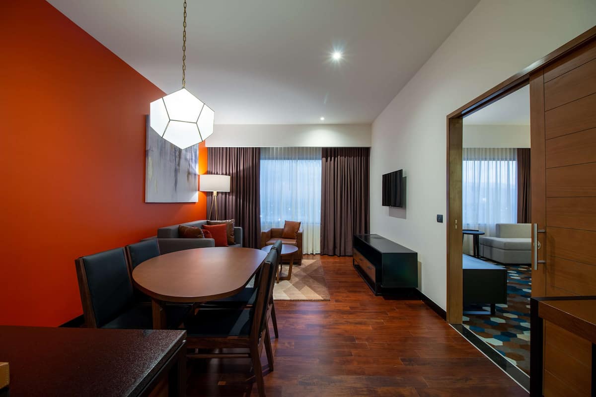 Quarto de hotel moderno com área de refeições, sofá, televisão e um espelho grande.