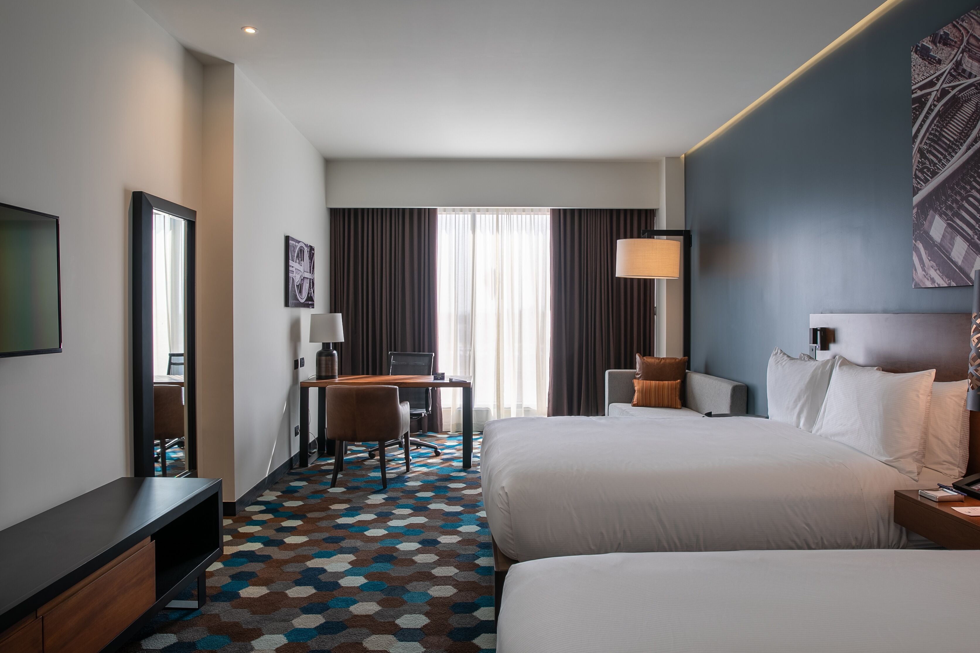 Habitación de hotel con dos camas, un escritorio con silla, una lámpara y un ventanal con cortinas.