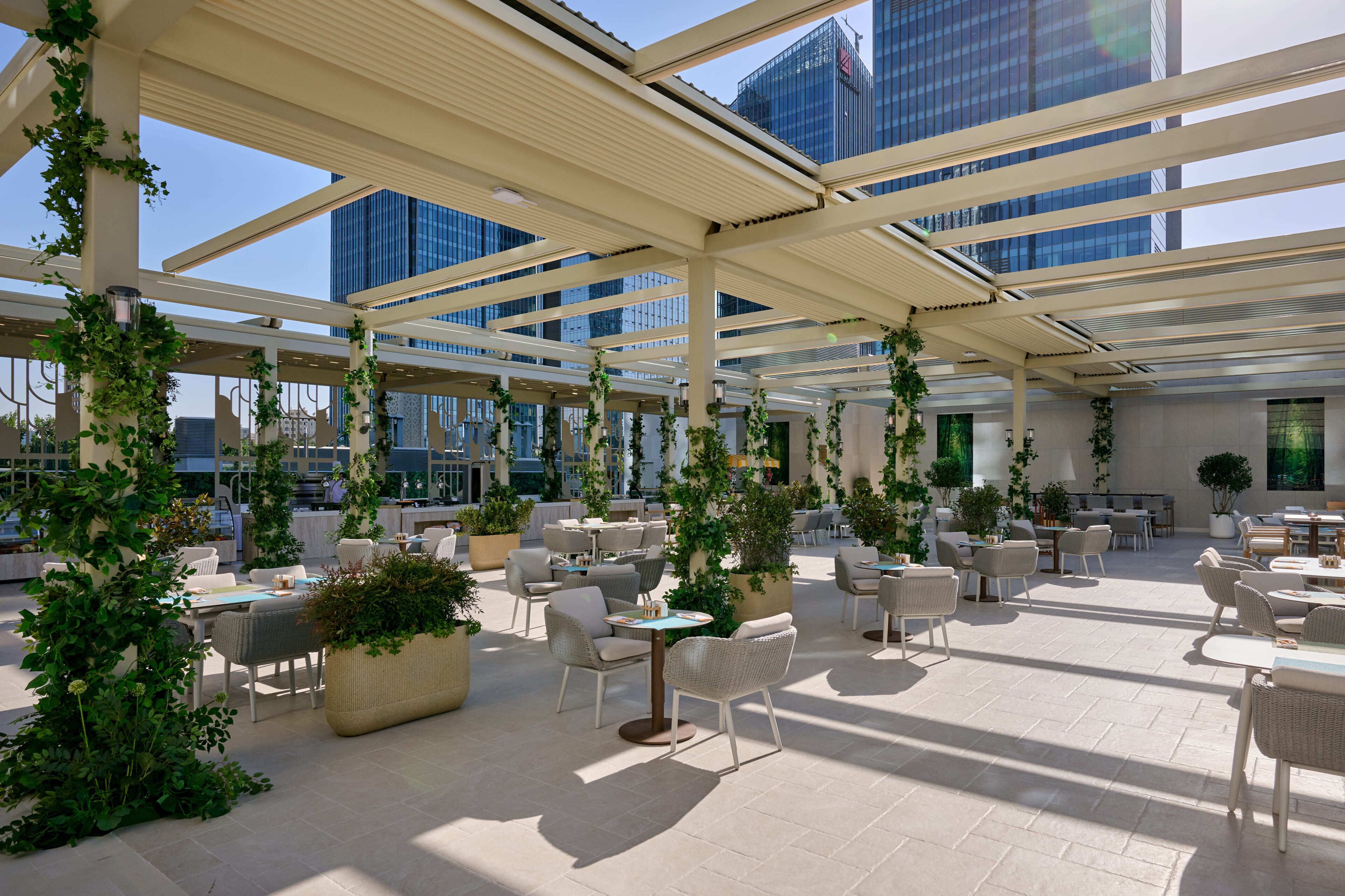 A spacious outdoor seating area with tables and chairs, surrounded by greenery and large windows offering views of the cityscape.