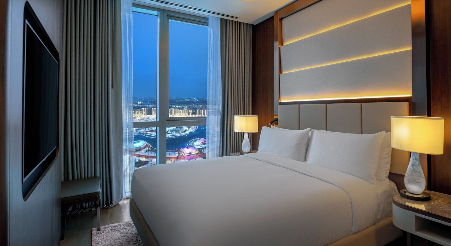 Habitación de hotel con una cama grande, dos lámparas de noche, un televisor y vistas de la ciudad a través de ventanales de gran altura.