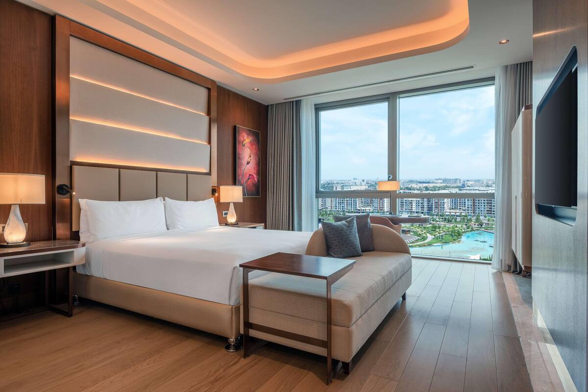 Una habitación de hotel moderna con una cama grande, un sofá, un televisor y vistas a un paisaje urbano y una piscina.