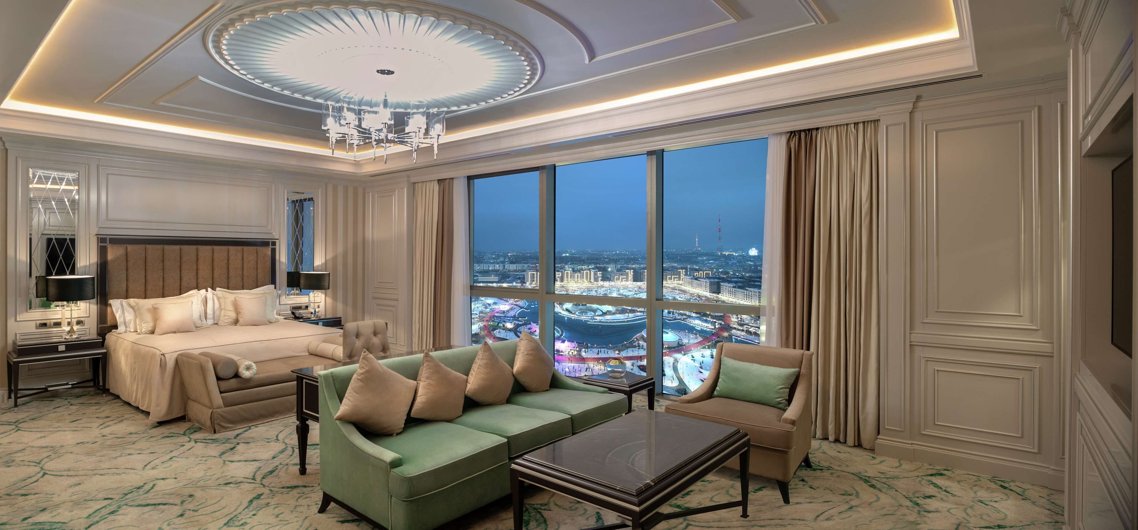 A spacious living room with a large window offering a city view, a green sofa, a beige sofa, a small table, and a chandelier.