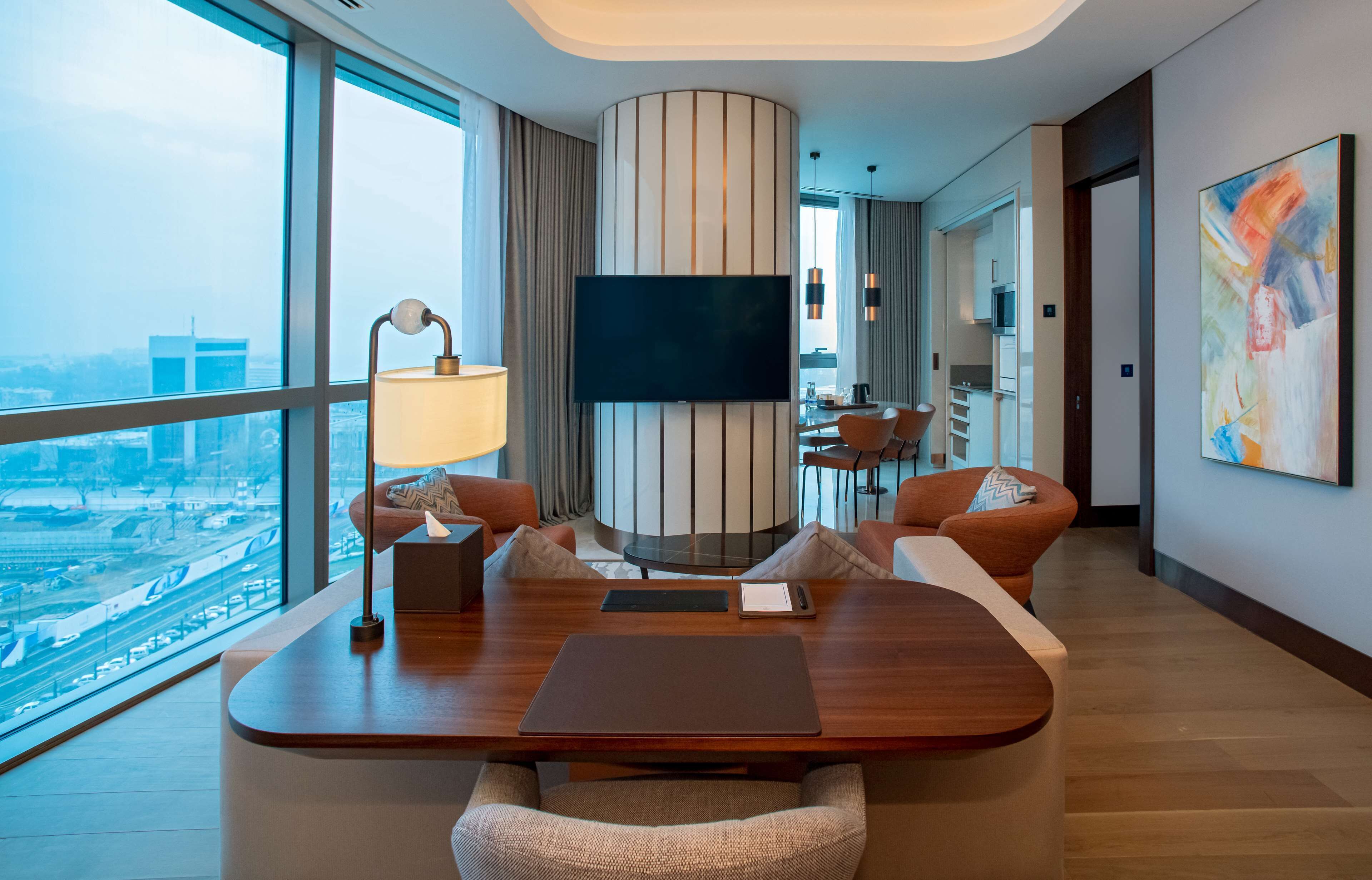 A modern hotel room with a large window offering a city view, a flat-screen TV mounted on a striped wall, a dining area with a table and chairs, and a contemporary art piece on the wall.