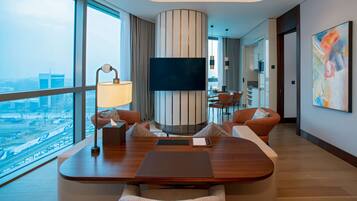 A modern hotel room with a large window offering a city view, a flat-screen TV mounted on a striped wall, a dining area with a table and chairs, and a contemporary art piece on the wall.