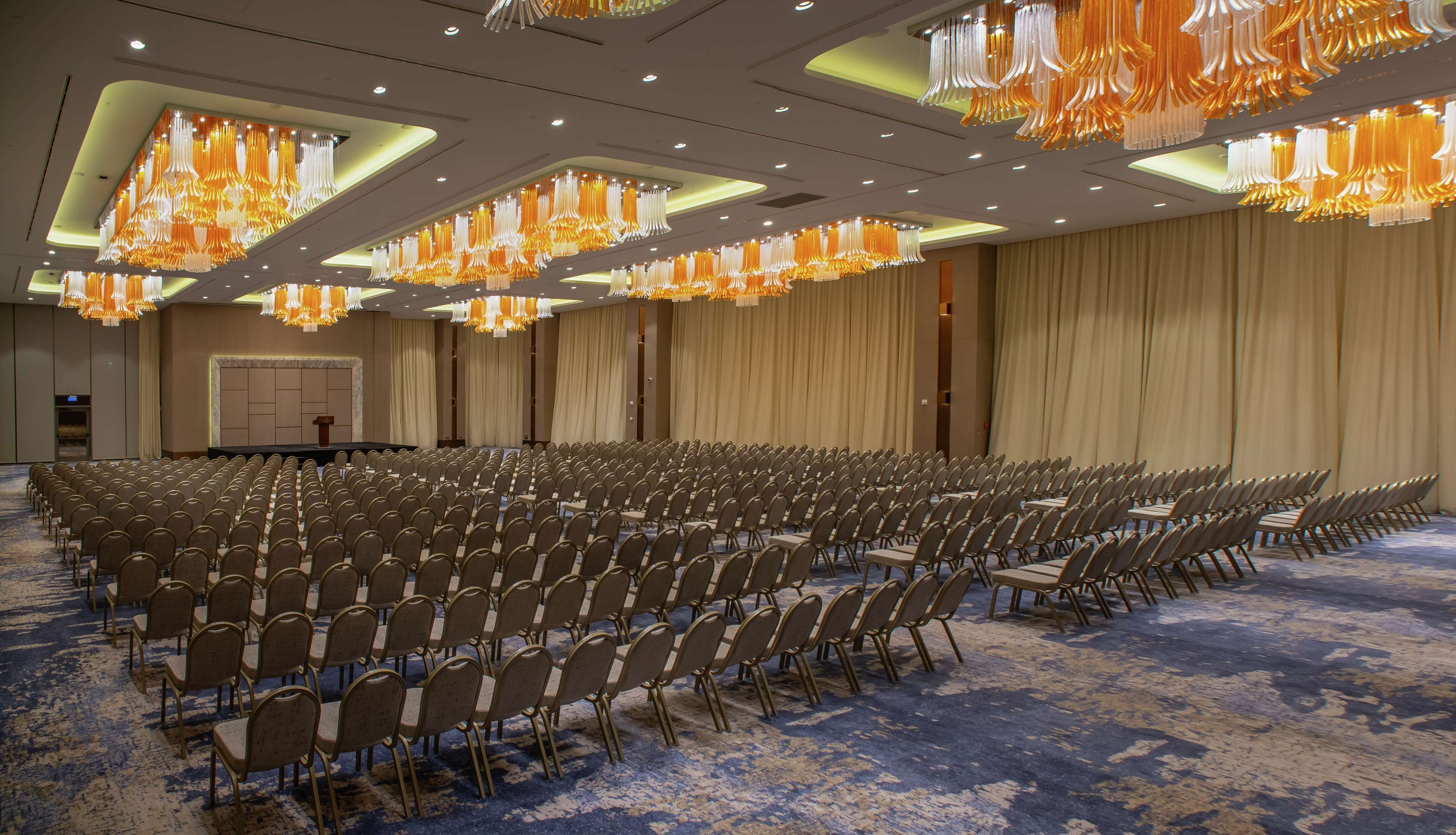 A large conference room with rows of chairs, a podium, and ornate chandeliers.