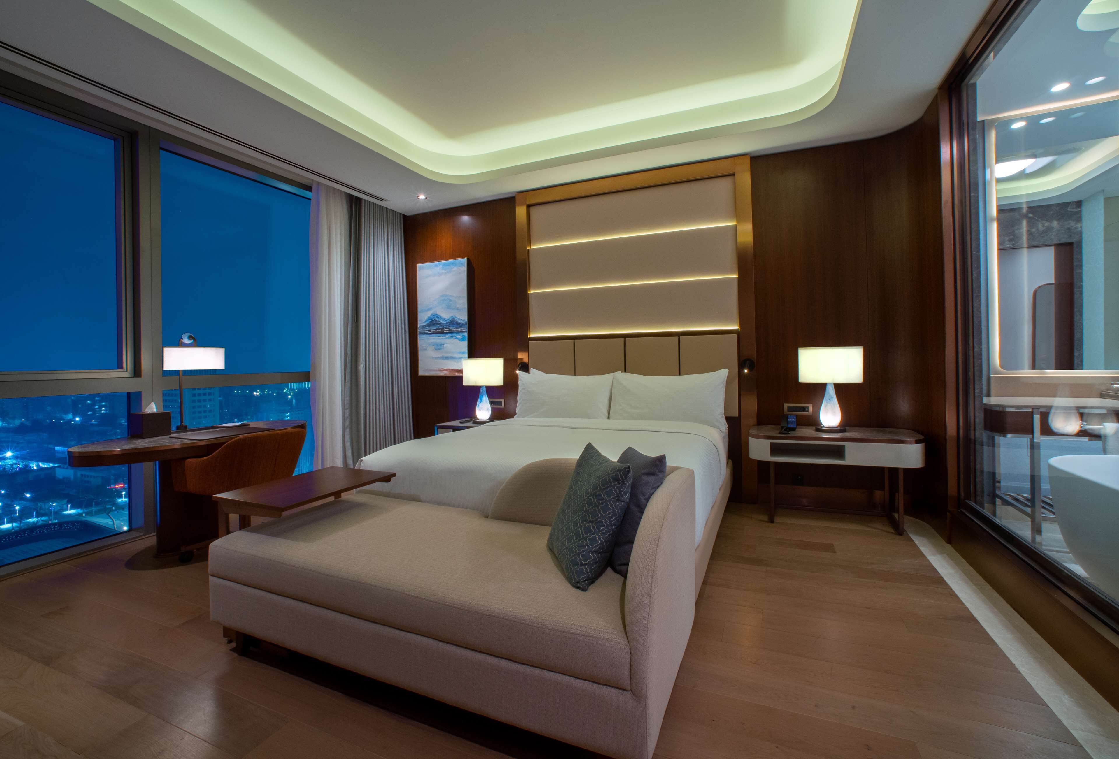 A modern hotel room with a large bed, a desk, and a view of the city at night.